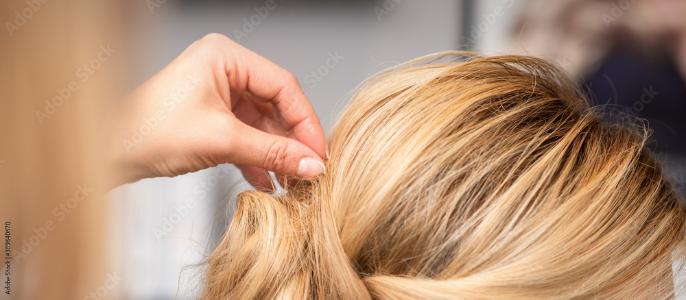 Naklejka premium Close up back view of hand of female hairdresser makes hairstyle of young blonde woman in beauty salon