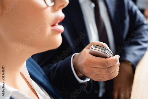 partial view of journalist with dictaphone interviewing politician during press conference, blurred foreground