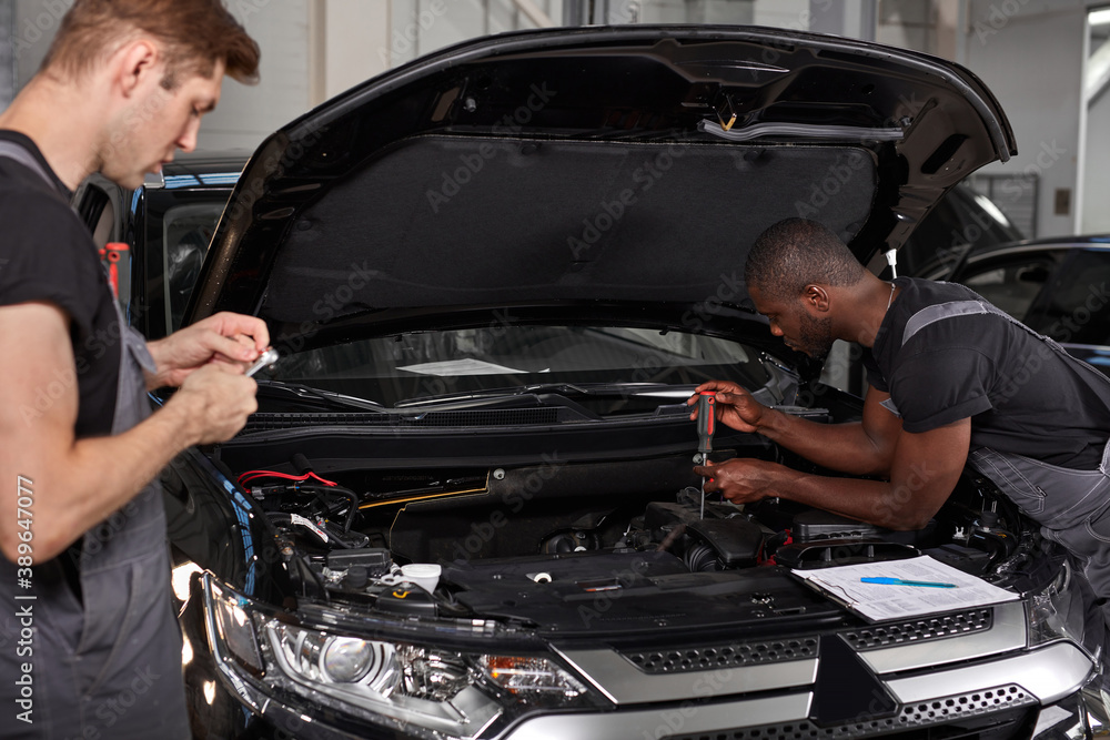 multi-ethnic team of african and caucasian men working in auto service together, friendly men in uniform repair the car hood multi-ethnic team of african and caucasian men working in auto service together, friendly men in uniform repair the car hood