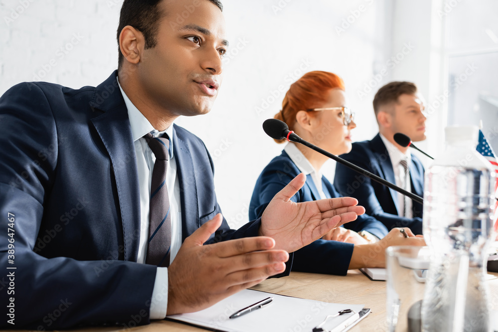 Obraz premium Indian politician gesturing, while speaking in microphone in boardroom with blurred colleagues on foreground