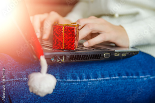 Woman in blue jeans and a white sweater holds a laptop with a Santa Claus hat and a red gift box on her lap and types. Concept of Christmas, New year, holiday