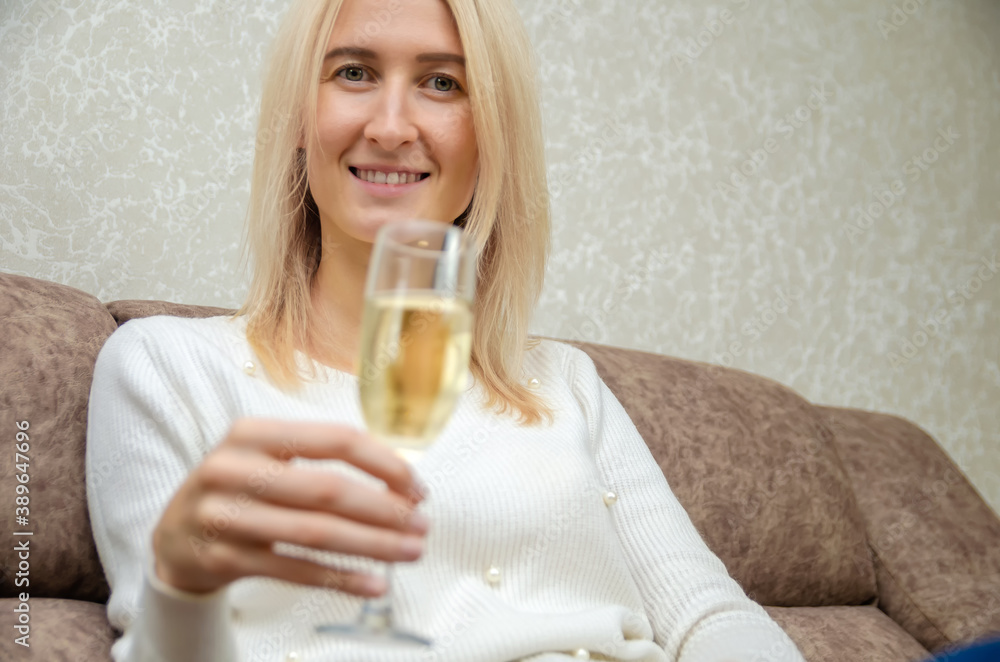 Young blonde woman in a white sweater with a glass of champagne is sitting on the sofa, copying the space. Young blonde woman in a white sweater with a glass of champagne is sitting on the sofa, copying the space.