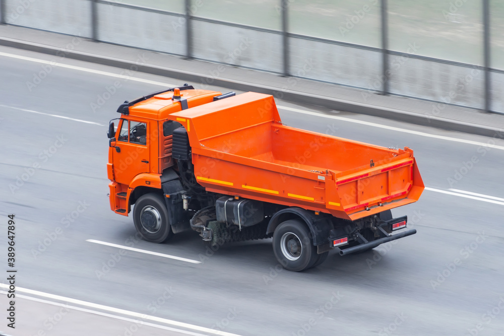 An orange dump truck with an empty body and a road cleaning brush drives on the highway.