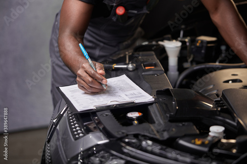 african professional auto service technician in uniform standing near car hood repairing and using check list for car inspect, indoors