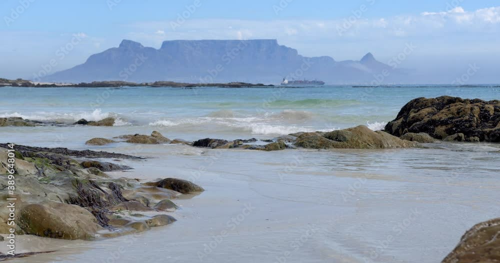 Waves gently splash over rocks in foreground with table mountain and a ship in the distance Waves gently splash over rocks in foreground with table mountain and a ship in the distance