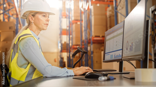Beautiful Female Worker Wearing Hard Hat Works on Personal Computer Counting Stock in the Retail Warehouse full of Shelves with Goods. Commerce, Distribution, Logistics