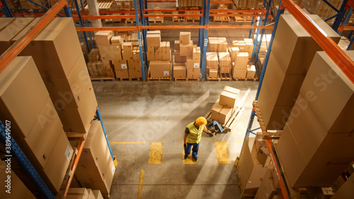 Top-Down View: Worker Moves Cardboard Boxes using Manual Pallet Truck, Walking between Rows of Shelves with Goods in Retail Warehouse. People Work in Product Distribution Logistics Center
