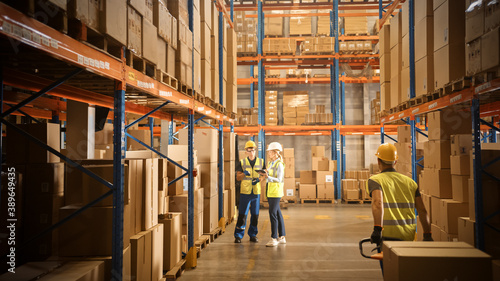 Retail Warehouse full of Shelves with Goods in Cardboard Boxes, Male and Female Managers Use Digital Tablet, Talk about Product Delivery. In Background Forklift Working in Logistics Storehouse photo