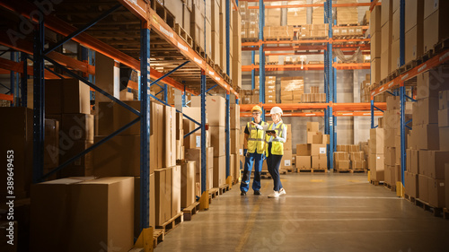 Retail Warehouse full of Shelves with Goods in Cardboard Boxes, Male and Female Managers Use Digital Tablet, Talk about Product Delivery. In Background Forklift Working in Logistics Storehouse