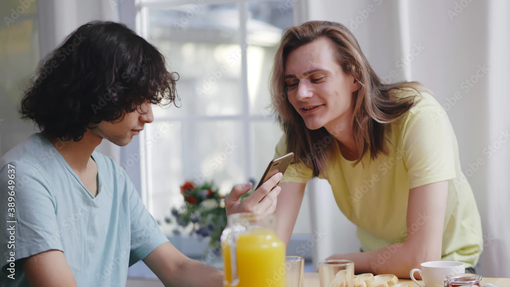 Gay couple eating breakfast and using smartphone at home in morning Gay couple eating breakfast and using smartphone at home in morning