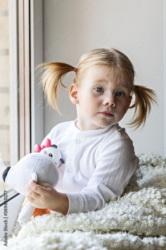 A little girl in white clothes plays with soft toys by the window. Self-isolation and quarantine content during the coronavirus pandemic in the world. A little girl in white clothes plays with soft toys by the window. Self-isolation and quarantine content during the coronavirus pandemic in the world.