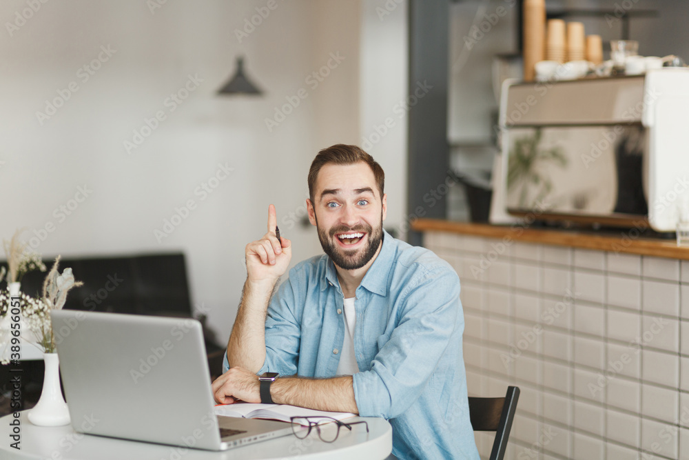 Excited young man sit alone at table in coffee shop cafe restaurant indoor working studying on laptop pc computer holding index finger up with great new idea. Freelance mobile office business concept. Excited young man sit alone at table in coffee shop cafe restaurant indoor working studying on laptop pc computer holding index finger up with great new idea. Freelance mobile office business concept.