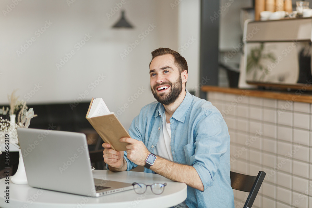 Cheerful man sitting alone at table in coffee shop cafe working or studying on laptop computer reading book relaxing in restaurant during free time indoors. Freelance mobile office business concept. Cheerful man sitting alone at table in coffee shop cafe working or studying on laptop computer reading book relaxing in restaurant during free time indoors. Freelance mobile office business concept.