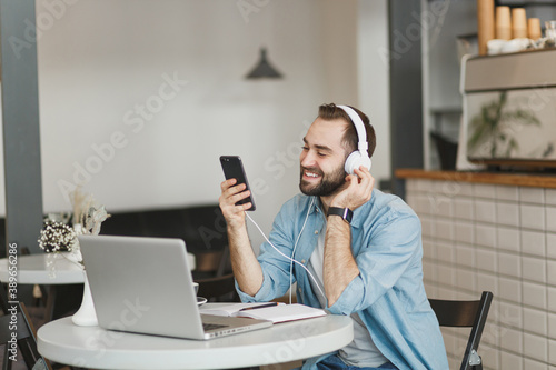 Smiling young man sitting at table in coffee shop cafe restaurant indoors working studying on laptop pc computer listening music with headphones cell phone. Freelance mobile office business concept.