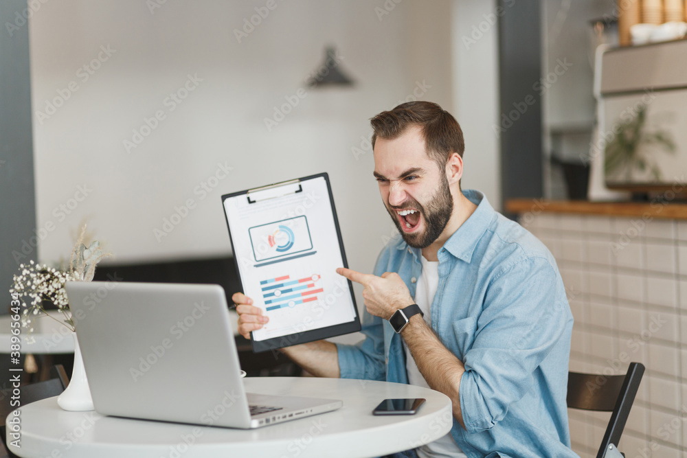 Angry young man sit at table in coffee shop cafe restaurant indoors working on laptop pc computer point on clipboard with papers document screaming swearing. Freelance mobile office business concept. Angry young man sit at table in coffee shop cafe restaurant indoors working on laptop pc computer point on clipboard with papers document screaming swearing. Freelance mobile office business concept.
