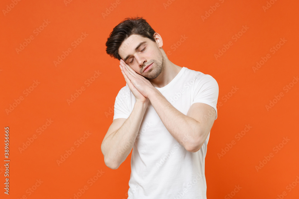 Relaxed handsome young man 20s wearing basic casual white blank empty t-shirt sleep with folded hands under cheek keeping eyes closed isolated on bright orange colour wall background, studio portrait.
