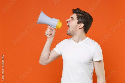 Displeased dissatisfied irritated young man 20s wearing basic casual empty white t-shirt standing screaming in megaphone looking aside isolated on bright orange colour background studio portrait.