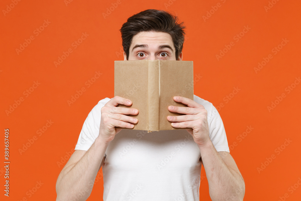 Shocked amazed young man 20s wearing basic casual white blank empty t-shirt standing reading covering face with book hiding looking camera isolated on bright orange colour background, studio portrait. Shocked amazed young man 20s wearing basic casual white blank empty t-shirt standing reading covering face with book hiding looking camera isolated on bright orange colour background, studio portrait.
