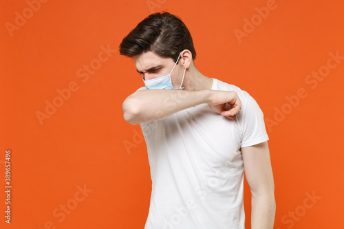 Sick young man in white t-shirt sterile face mask to safe from coronavirus virus covid-19 during quarantine coughing or sneezing covering mouth with hand isolated on orange background studio portrait. © ViDi Studio