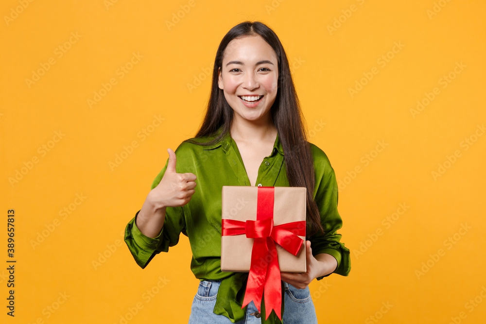 Funny young brunette asian woman in basic green shirt standing hold red present box with gift ribbon bow showing thumb up looking camera isolated on bright yellow colour background, studio portrait. Funny young brunette asian woman in basic green shirt standing hold red present box with gift ribbon bow showing thumb up looking camera isolated on bright yellow colour background, studio portrait.