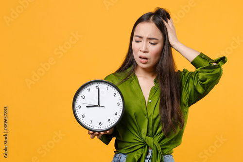 Concerned shocked worried young brunette asian woman 20s wearing basic green shirt standing hold clock put hand on head looking camera isolated on bright yellow colour background studio portrait.