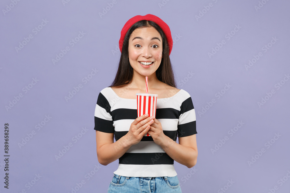 Smiling excited young brunette asian woman 20s wearing striped t-shirt red beret standing hold plastic cup of cola or soda looking camera isolated on pastel violet colour background studio portrait.