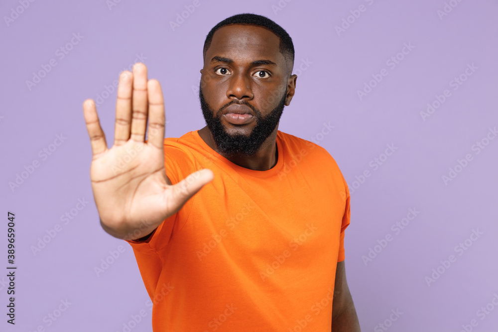 Serious displeased young african american man wearing basic casual orange t-shirt standing showing stop gesture with palm looking camera isolated on pastel violet background studio portrait. Serious displeased young african american man wearing basic casual orange t-shirt standing showing stop gesture with palm looking camera isolated on pastel violet background studio portrait.