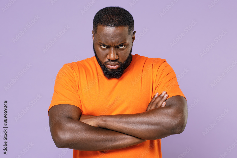 Dissatisfied displeased young african american man in basic casual blank empty orange t-shirt standing holding hands crossed looking camera isolated on pastel violet colour background studio portrait. Dissatisfied displeased young african american man in basic casual blank empty orange t-shirt standing holding hands crossed looking camera isolated on pastel violet colour background studio portrait.