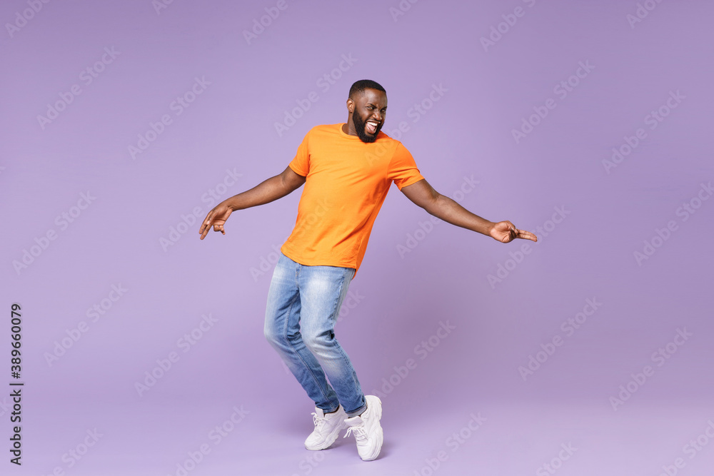 Full length of excited funny young african american man 20s in basic casual orange t-shirt dancing standing on toes pointing index fingers aside isolated on pastel violet background studio portrait.