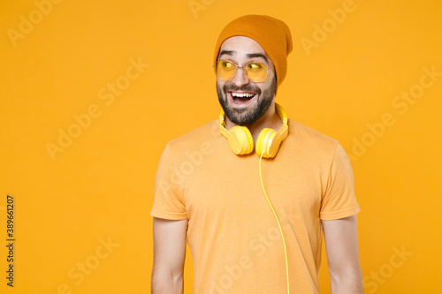 Excited surprised young bearded man 20s wearing basic casual t-shirt headphones eyeglasses hat standing keeping mouth open looking aside isolated on bright yellow colour background, studio portrait.