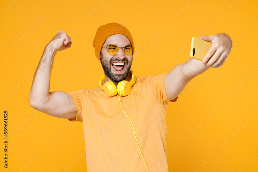 Happy joyful young bearded man wearing basic casual t-shirt headphones eyeglasses hat standing doing selfie shot on mobile phone doing winner gesture isolated on yellow background, studio portrait.