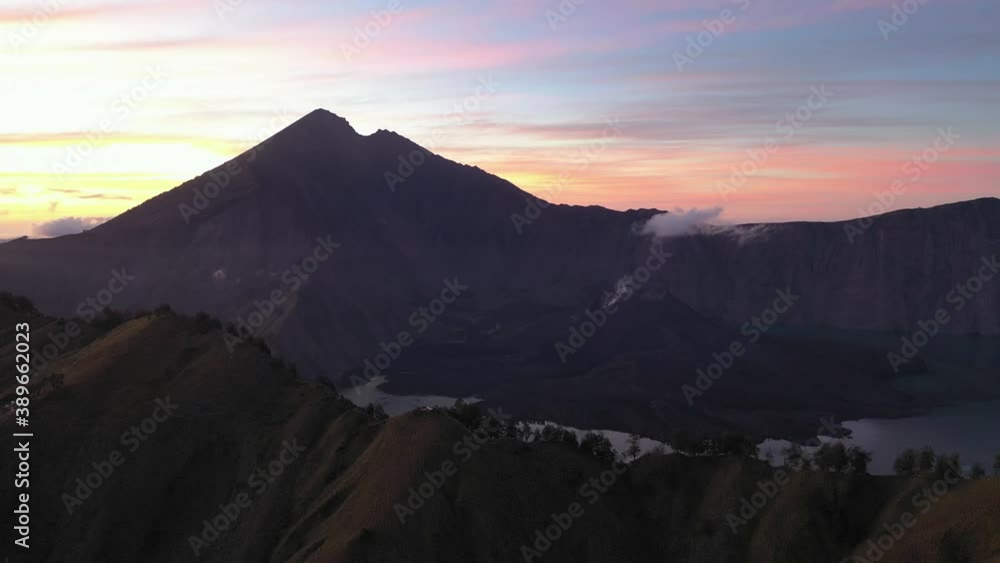 Cinematic aerial upwards reveal of Rinjani Lake and summit at predawn, thus a colourful dramatic sky with dark foreboding foreground. Lombok Island, Indonesia. Cinematic aerial upwards reveal of Rinjani Lake and summit at predawn, thus a colourful dramatic sky with dark foreboding foreground. Lombok Island, Indonesia.