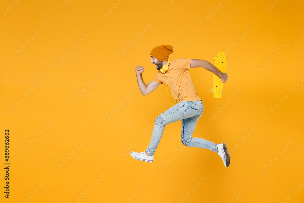 Full length side view of cheerful funny young man 20s wearing basic casual t-shirt headphones hat jumping like running hold skateboard isolated on bright yellow colour background, studio portrait.