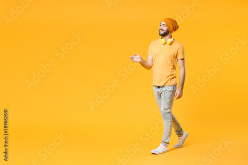 Full length side view of smiling young man wearing basic casual t-shirt headphones hat standing with outstretched hand for greeting looking aside isolated on bright yellow background, studio portrait.