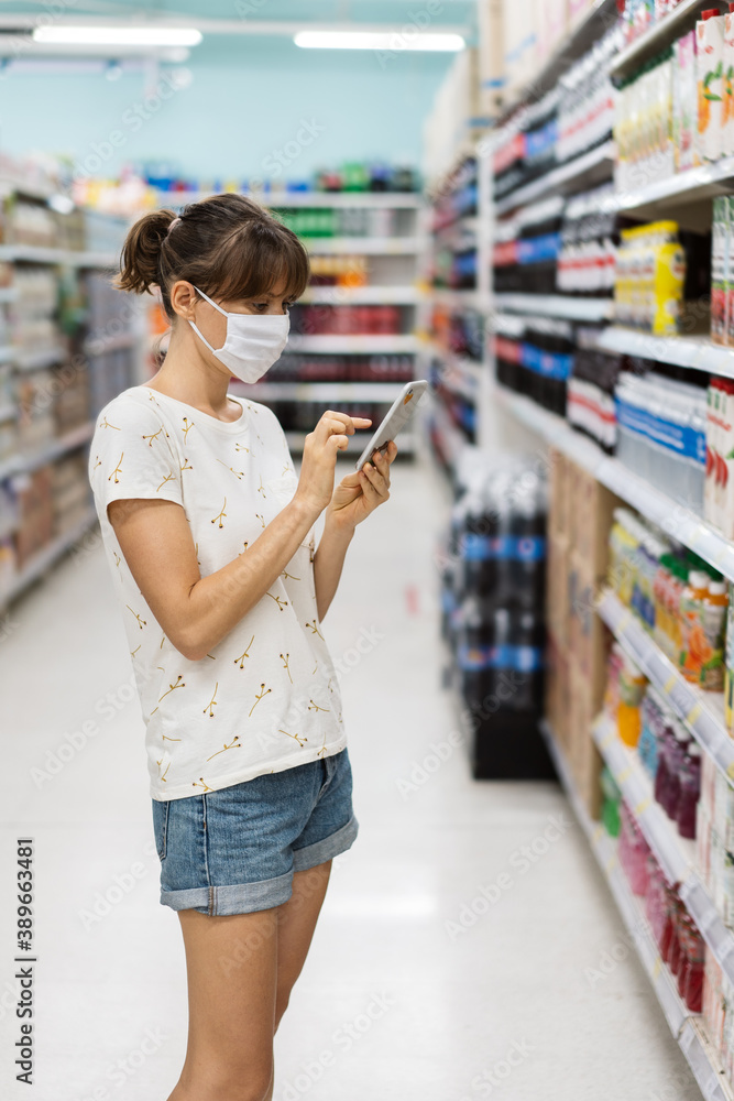 Young woman with face mask using mobile phone and buying groceries in the supermarket
