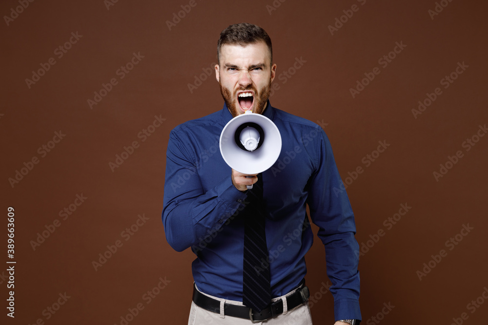 Angry irritated bearded young business man wearing blue shirt tie screaming in megaphone looking camera isolated on brown colour background studio portrait. Achievement career wealth business concept. Angry irritated bearded young business man wearing blue shirt tie screaming in megaphone looking camera isolated on brown colour background studio portrait. Achievement career wealth business concept.