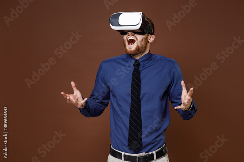 Shocked bearded young business man wearing blue shirt tie watching in vr headset gadget spreading hands isolated on brown colour background studio portrait. Achievement career wealth business concept.