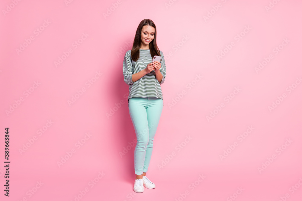 Full size photo of optimistic cute young woman stand write sms wear blue pants sneakers shirt isolated on pastel pink color background Full size photo of optimistic cute young woman stand write sms wear blue pants sneakers shirt isolated on pastel pink color background