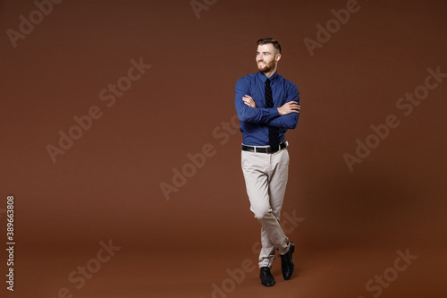 Full length of smiling successful young business man in blue shirt tie holding hands crossed looking aside isolated on brown background studio portrait. Achievement career wealth business concept.