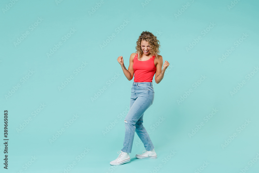 Full length side view of joyful young blonde woman 20s in pink tank top jeans standing clenching fists doing winner gesture keeping eyes closed isolated on blue turquoise background, studio portrait. Full length side view of joyful young blonde woman 20s in pink tank top jeans standing clenching fists doing winner gesture keeping eyes closed isolated on blue turquoise background, studio portrait.