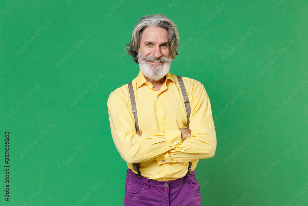 Smiling cheerful elderly gray-haired mustache bearded man in casual yellow shirt suspenders standing holding hands crossed looking camera isolated on bright green colour background, studio portrait.
