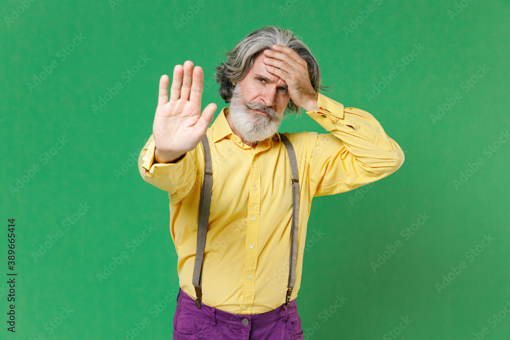 Dissatisfied elderly gray-haired mustache bearded man wearing casual yellow shirt suspenders showing STOP gesture with palm put hand on head isolated on bright green colour background studio portrait. Dissatisfied elderly gray-haired mustache bearded man wearing casual yellow shirt suspenders showing STOP gesture with palm put hand on head isolated on bright green colour background studio portrait.