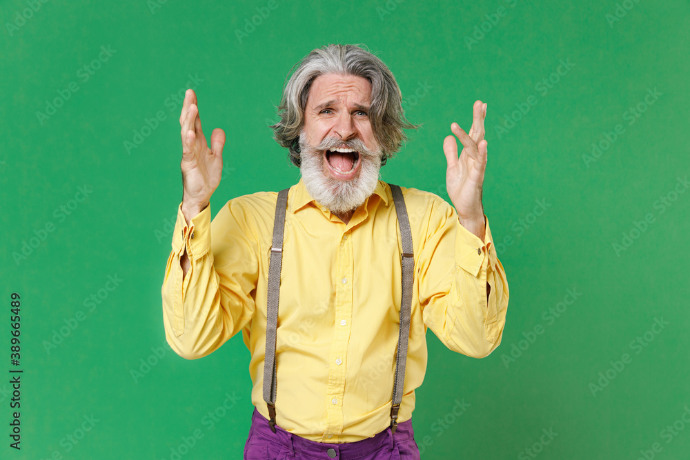 Angry irritated elderly gray-haired mustache bearded man wearing casual yellow shirt suspenders standing spreading hands screaming swearing isolated on bright green colour background, studio portrait.