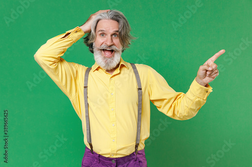 Excited elderly gray-haired mustache bearded man wearing basic yellow shirt suspenders put hand on head pointing index finger aside on mock up copy space isolated on green background studio portrait. © ViDi Studio