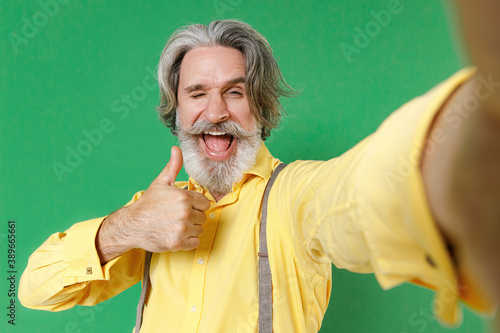 Close up of blinking elderly gray-haired mustache bearded man wearing casual yellow shirt suspenders doing selfie shot on mobile phone showing thumb up isolated on green background studio portrait. © ViDi Studio