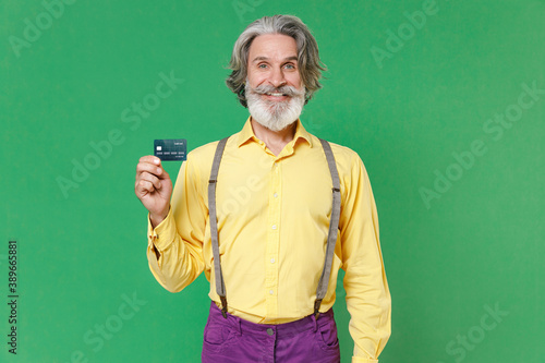 Smiling funny elderly gray-haired mustache bearded man wearing casual yellow shirt suspenders standing hold credit bank card looking camera isolated on bright green colour background studio portrait.