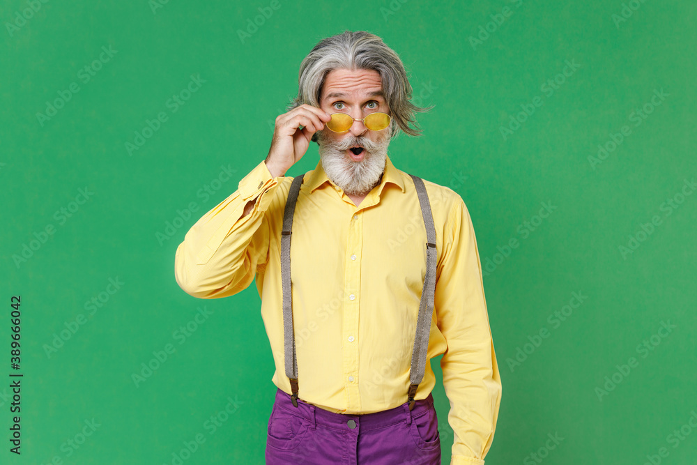 Shocked amazed elderly gray-haired mustache bearded man wearing casual yellow shirt suspenders eyeglasses keeping mouth open looking camera isolated on bright green colour background, studio portrait. Shocked amazed elderly gray-haired mustache bearded man wearing casual yellow shirt suspenders eyeglasses keeping mouth open looking camera isolated on bright green colour background, studio portrait.