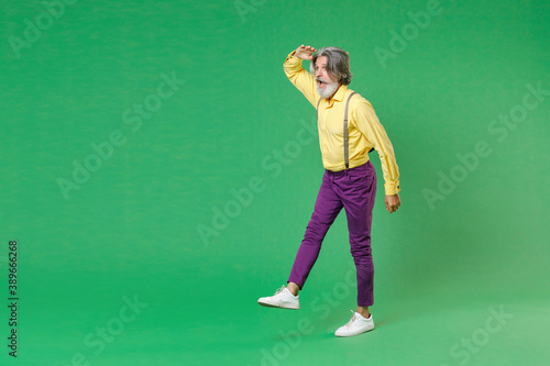 Full length side view of shocked elderly gray-haired mustache bearded man in yellow shirt suspenders holding hand at forehead looking far away distance isolated on green background, studio portrait. © ViDi Studio