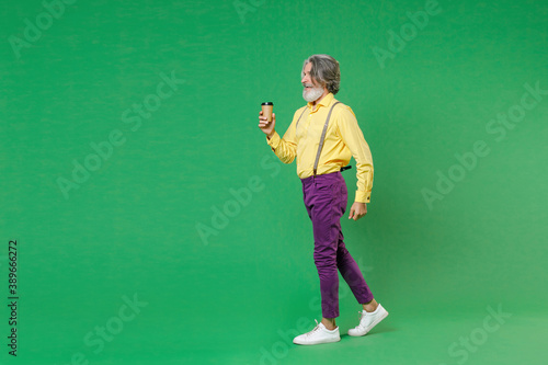 Full length side view of elderly gray-haired mustache bearded man wearing casual yellow shirt suspenders hold paper cup of coffee or tea isolated on bright green colour background, studio portrait. © ViDi Studio