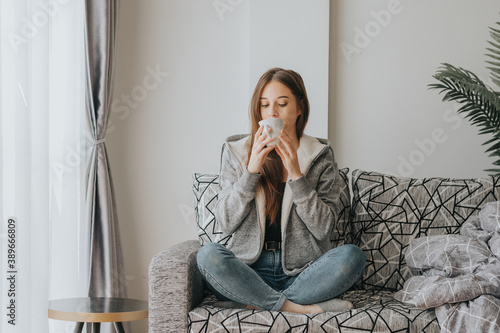 Фотография Young Woman sitting on sofa wearing sweater sipping coffee or tea from white mug